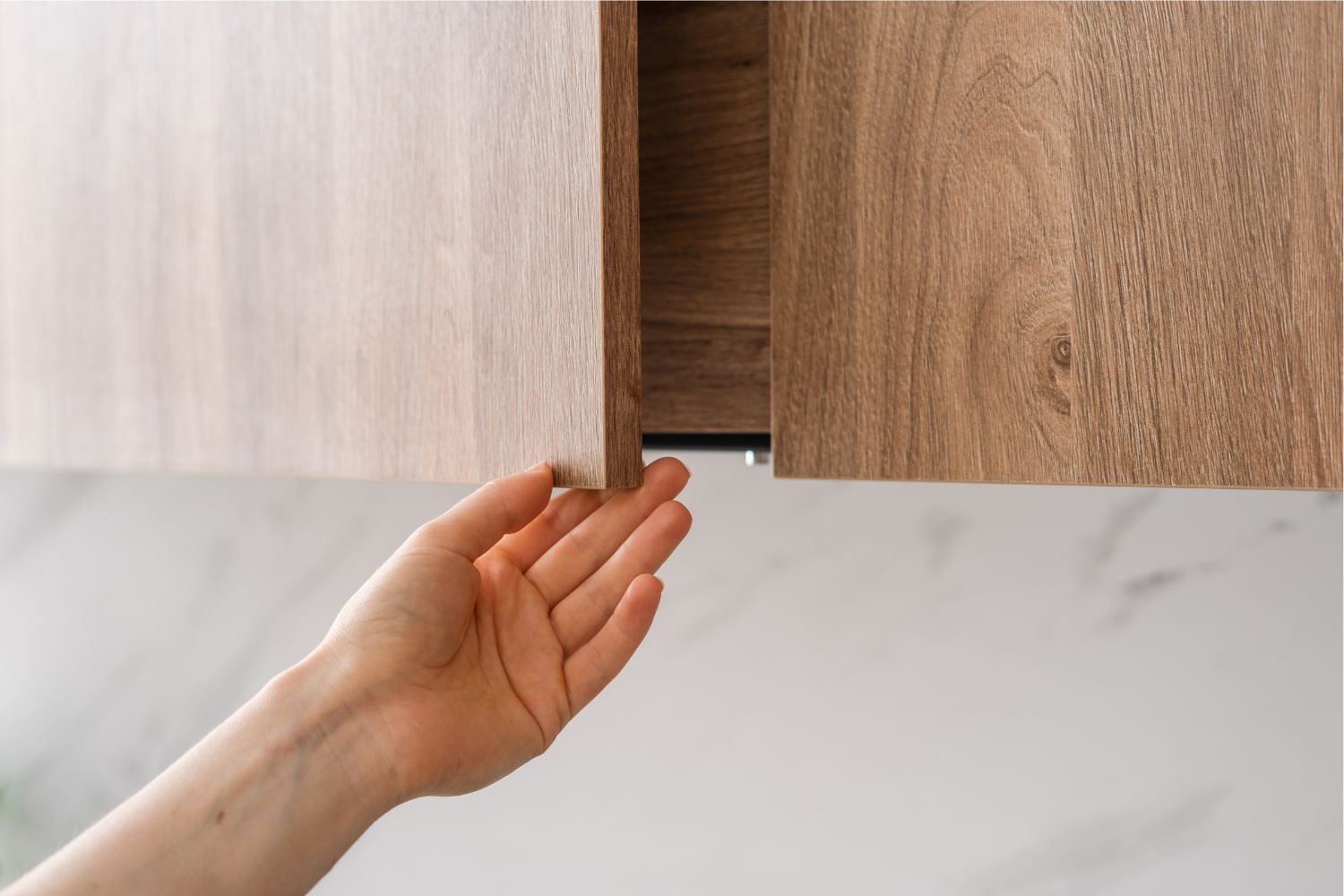 Hand opening a light brown kitchen cabinet. White wall in the background.